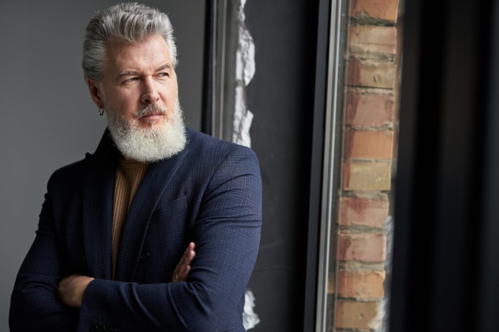 Calm gray haired middle aged man in business casual wear looking out a window while standing in loft